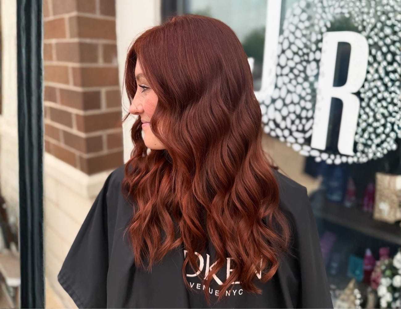 Red-haired person with wavy style in front of a brick wall and salon window.