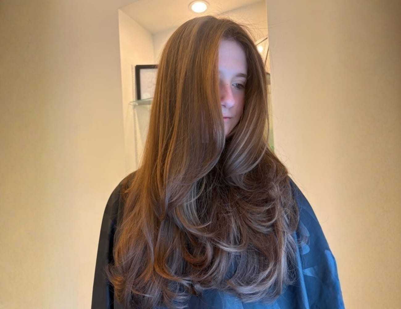 Young person with long, flowing brown hair stands against a light background.